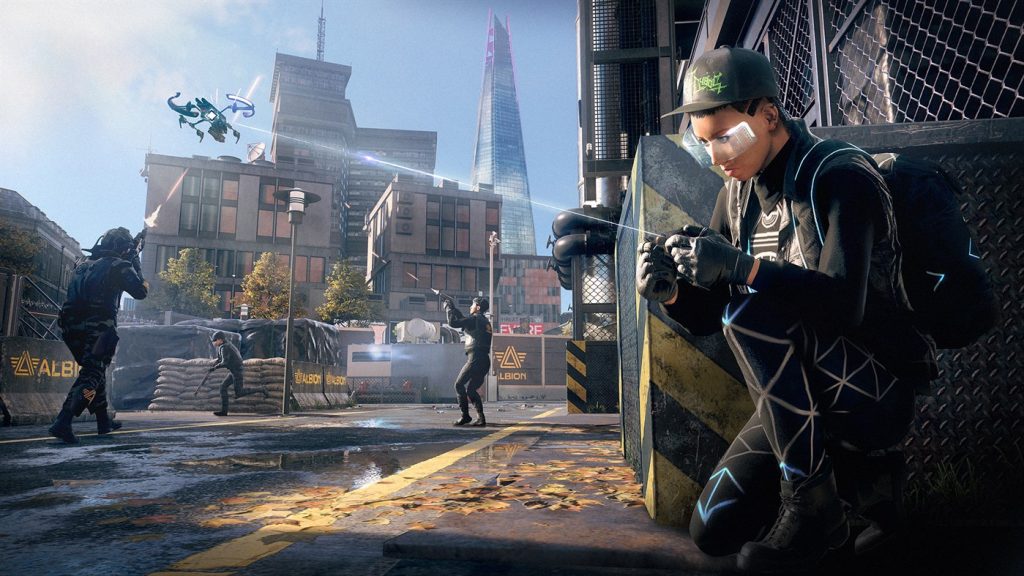 A gameplay image from Watch Dogs: Legion. A realistic image of a street battle in London. In the background, three people in black riot gear are trying to shoot down a drone. In the foreground, a woman in a black baseball hat and futuristic clothing, including leggings with a glowing triangle pattern, crouches behind a building. She has a holographic screen projected in front of her eyes and is holding a controller in her hands; a white line connects her controller to the drone. 
