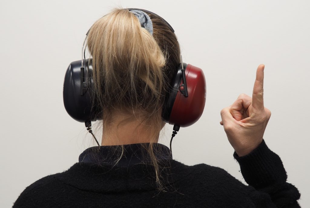 An image of a person taking a hearing test. The person is wearing a set of earphones- one ear is black, and the other is red- and pointing up with one finger.
