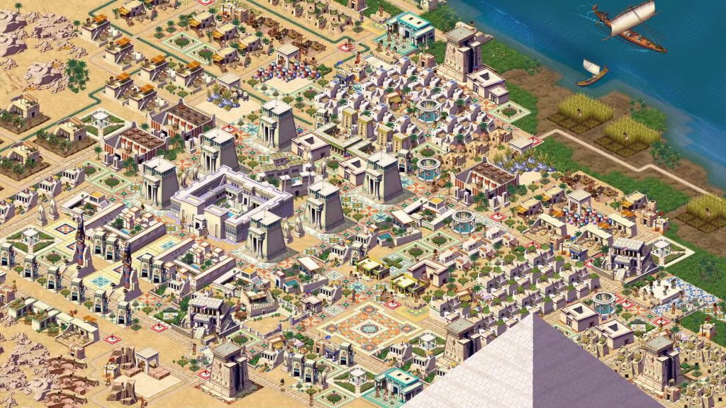 A stylized, isometric depiction of an Egyptian city from Pharaoh: A New Era. The city is in the desert, with many small houses at the edges and larger, more imposing buildings in the center. To the top right of the image is a riverbank covered with fields. Above the fields is the dark blue river. Two boats float in the water. At the bottom right corner of the image, the tip of a pyramid covers everything behind it.