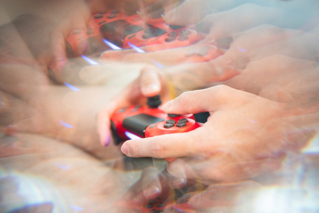 A distorted photograph of a pale-skinned person's hands on a red Playstation controller. The photograph is layered over itself to create a ghostly or blurry effect. 